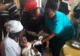 Vaccination of the first child by a healthcare worker, assisted by Amb. Julie Endee during the MCV2 launch in Bomi County, Liberia