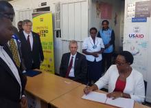 Health CS Kariuki signs a visitors’ book when she visited Mama Lucy Kibaki Hospital in commemoration of World TB Day. She was accompanied by MOH officials as well as Mr Robert Godec (seated) US Ambassador to Kenya and Dr Rudi Eggers, WHO Representative, Kenya 