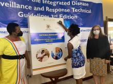 WHO'S Surveillance Officer Dr Solome Okware signs onto the dummy at the launch as State Minister of Health in Charge of General Duties, Hon Anifa Kawooya and CDC Country Director, Dr Lisa Nelson look on.