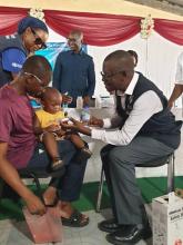 Dr. Momodou T. Nyassi (standing) shares a light moment with a parent during the launch of the 2025 Measles–Rubella Campaign, joined by Dr. Nathan Bakyaita, WHO Representative in The Gambia and Dr. Oumie Jagne, Global Polio Eradication Coordinator, WHO The Gambia. 