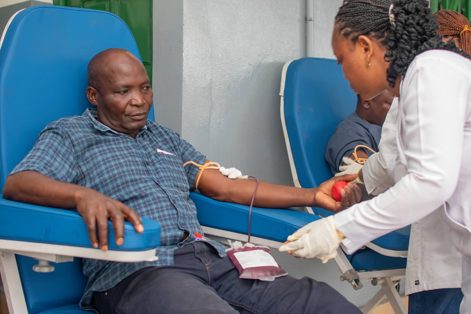 A donor reclines in a blue donation chair while a health worker hands him a heart-shaped stress ball